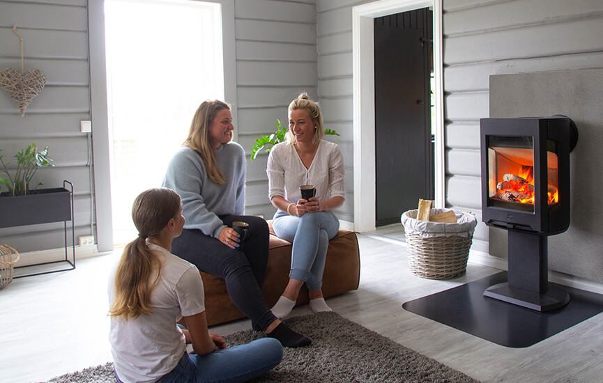 Trzy dziewczyny siedzą na brązowej pufie w szarym salonie przed piecem na drewno Jøtul F 167